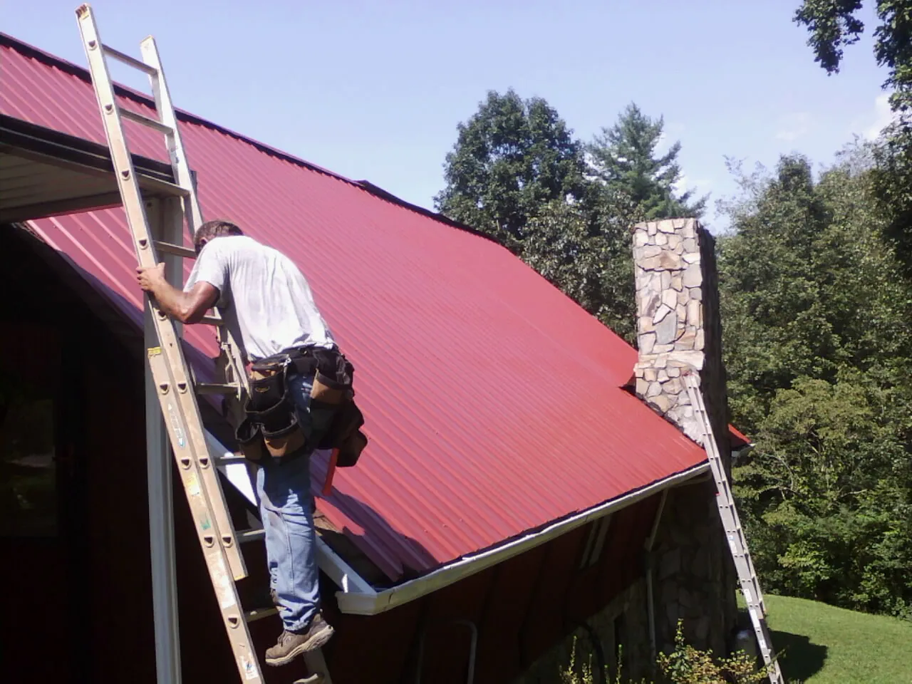 Roofer on a ladder beside a red metal roof and stone chimney — Soffit & Fascia Repair by Roofing Brothers in Parsons